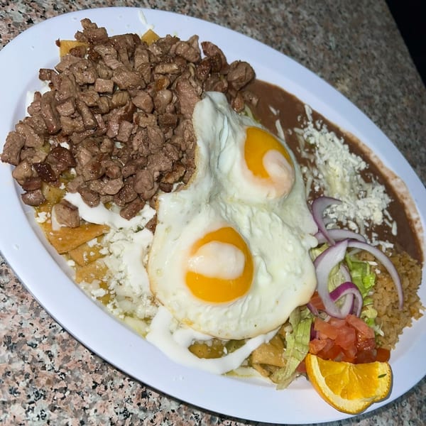 A plate of Mexican chilaquiles with steak, two fried eggs, refried beans, rice, and an orange slice.