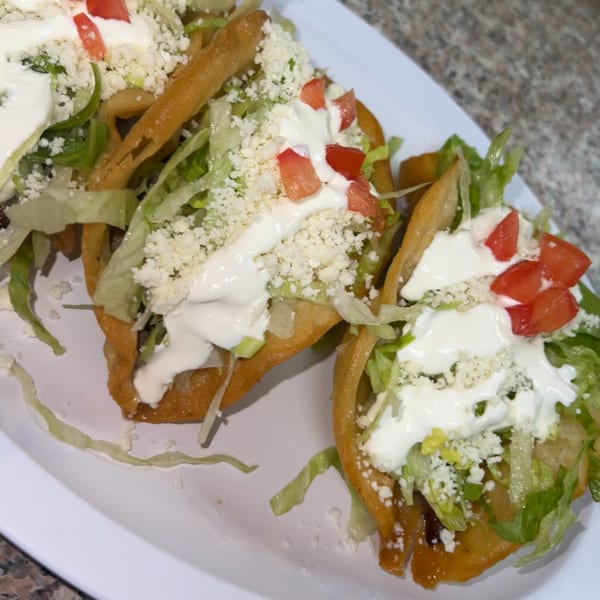 Three crispy Mexican tacos topped with lettuce, crumbled cheese, diced tomatoes, and sour cream.